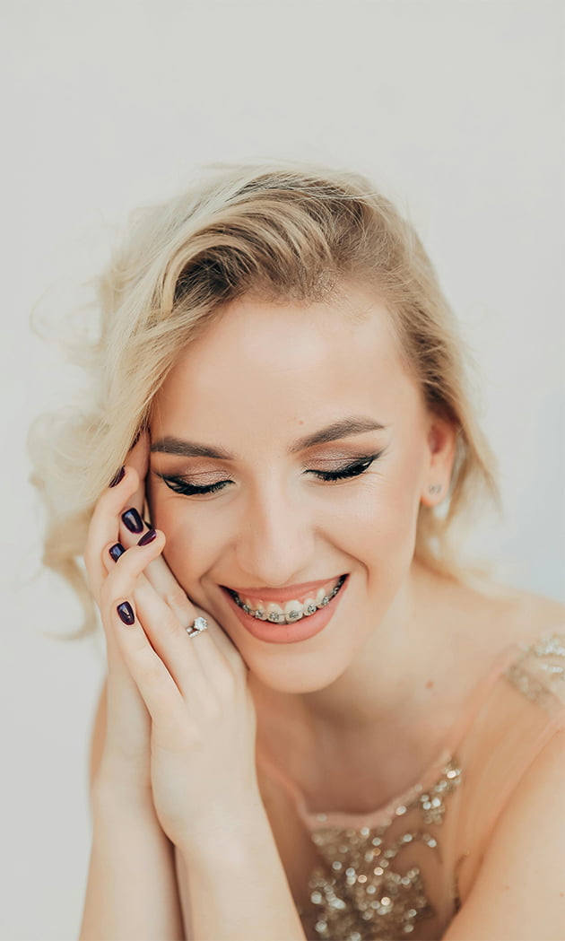 nasmješena žena svijetle kose prekriženih ruku sa aparatićem za odrasle u bež haljini sa sivom pozadinom